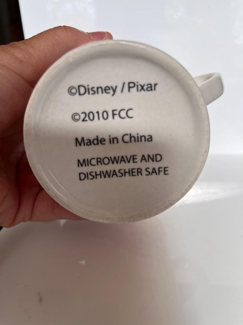 K&ouml;nnte beinhalten: Wei&szlig;er Keramikbecher mit Henkel. Der Boden des Bechers zeigt den Text "&copy;Disney / Pixar", "&copy;2010 FCC", "Made in China" und "MICROWAVE AND DISHWASHER SAFE" in schwarzer Schrift.