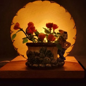 Könnte beinhalten: Eine Vintage-Lampe mit einem runden, cremefarbenen Schirm und einem Keramiksockel mit einem Blumenarrangement und einer Figur. Die Lampe ist eingeschaltet und strahlt ein warmes Licht aus.