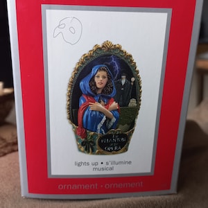 May include: A musical ornament featuring a scene from the Phantom of the Opera. The ornament depicts Christine Daaé in a blue gown, with the Phantom in the background. The ornament lights up and plays music. The text "The Phantom of the Opera" is printed on the box.