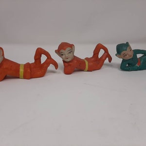 May include: Three ceramic figurines of elves, two are orange and one is green. The elves are lying on their stomachs with their arms outstretched.