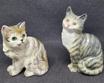 May include: Two ceramic cat figurines, one sitting upright and the other lying down. Both cats are grey and white with black stripes. The cat sitting upright has green eyes and a pink nose.
