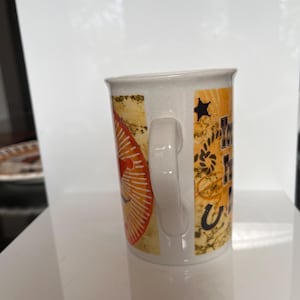 K&ouml;nnte beinhalten: Wei&szlig;er Keramikbecher mit Henkel. Der Becher hat ein Design mit orangefarbenen, gelben und schwarzen Elementen, darunter ein Stern, ein Hufeisen und Text. Der Becher steht auf einer wei&szlig;en Oberfl&auml;che.