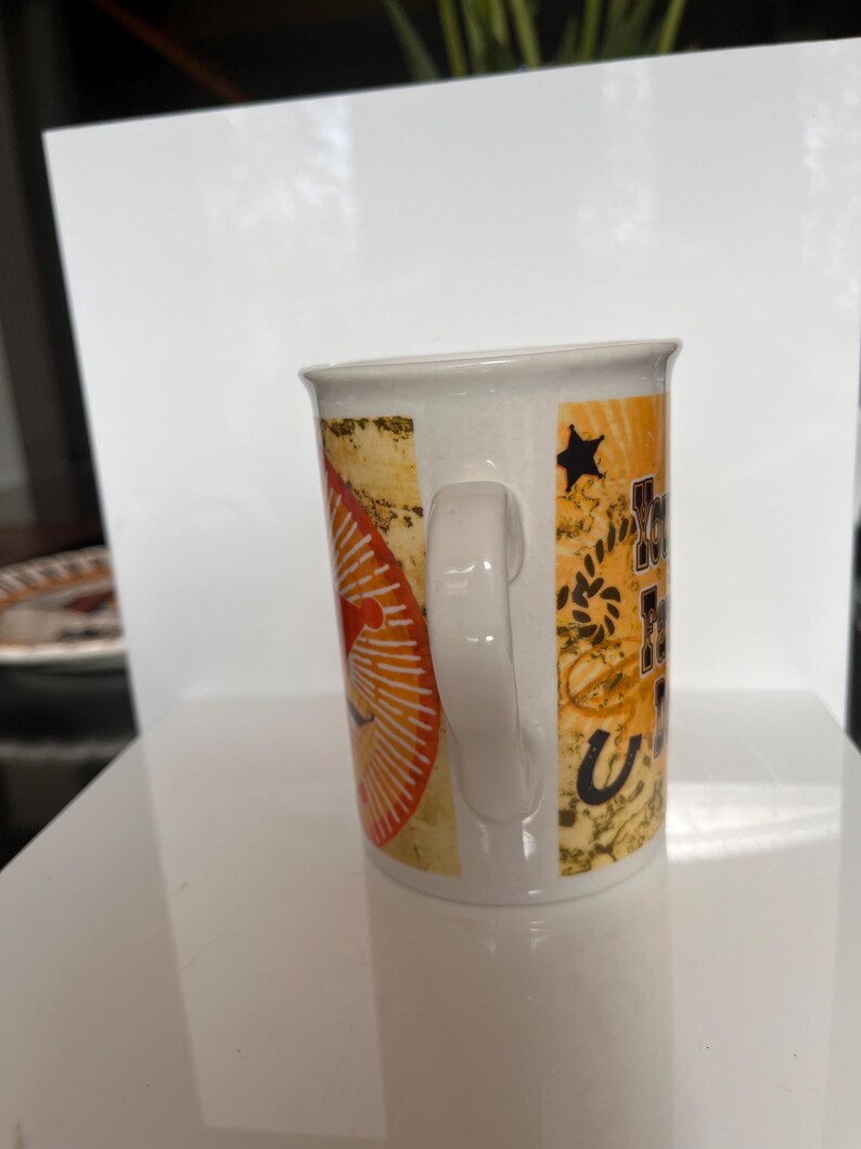 K&ouml;nnte beinhalten: Wei&szlig;er Keramikbecher mit Henkel. Der Becher hat ein Design mit orangefarbenen, gelben und schwarzen Elementen, darunter ein Stern, ein Hufeisen und Text. Der Becher steht auf einer wei&szlig;en Oberfl&auml;che.
