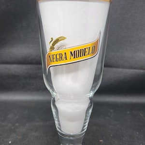 May include: A clear glass beer pilsner with a gold rim and the words "NEGRA MODELO" in a gold and black banner. The glass has a long stem and a wide, conical top. The glass is empty.