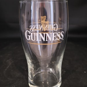 May include: A clear, tulip-shaped Guinness pint glass. The glass features the Guinness logo in white with a gold harp emblem. The glass is empty and sits on a dark surface.