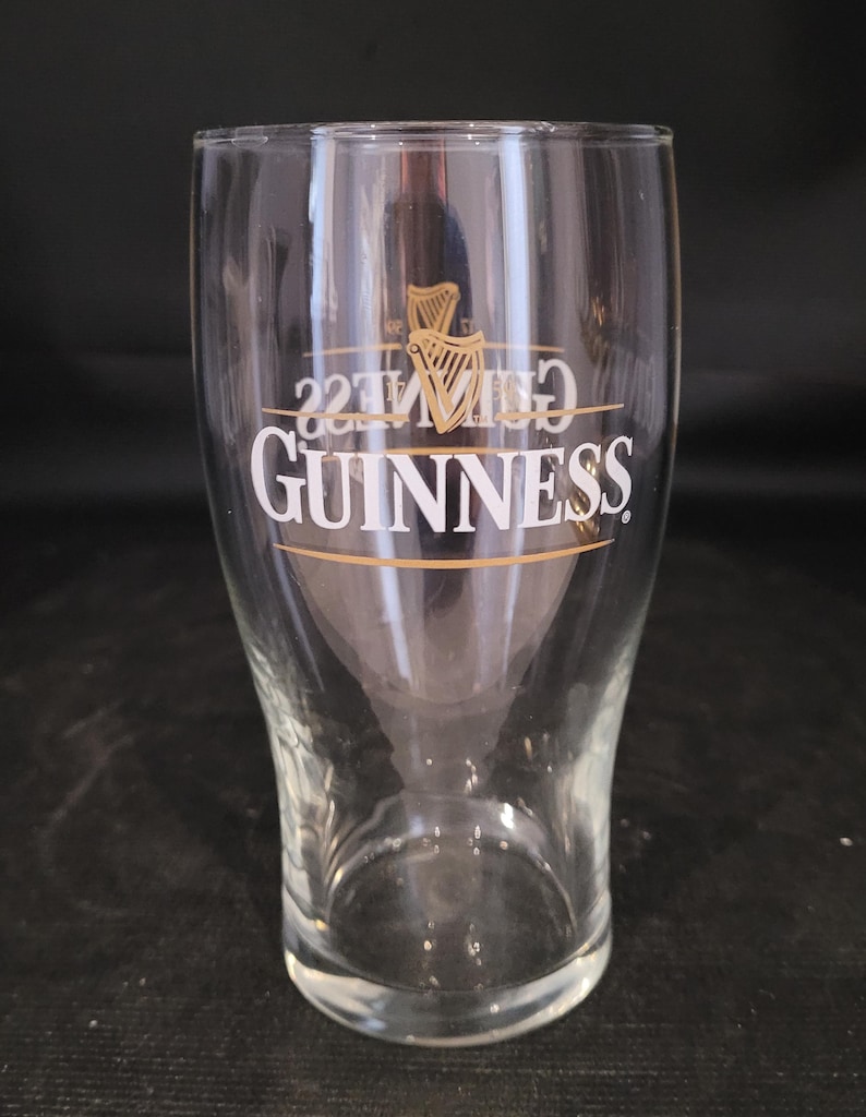 May include: A clear, tulip-shaped Guinness pint glass. The glass features the Guinness logo in white with a gold harp emblem. The glass is empty and sits on a dark surface.
