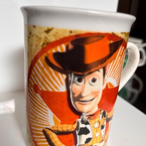 K&ouml;nnte beinhalten: Wei&szlig;er Keramikbecher mit einer farbenfrohen Illustration von Woody aus Toy Story. Das Design zeigt Woodys Gesicht, Hut und Outfit vor einem roten und orangefarbenen Hintergrund. Der Becher hat einen wei&szlig;en Henkel und Rand.