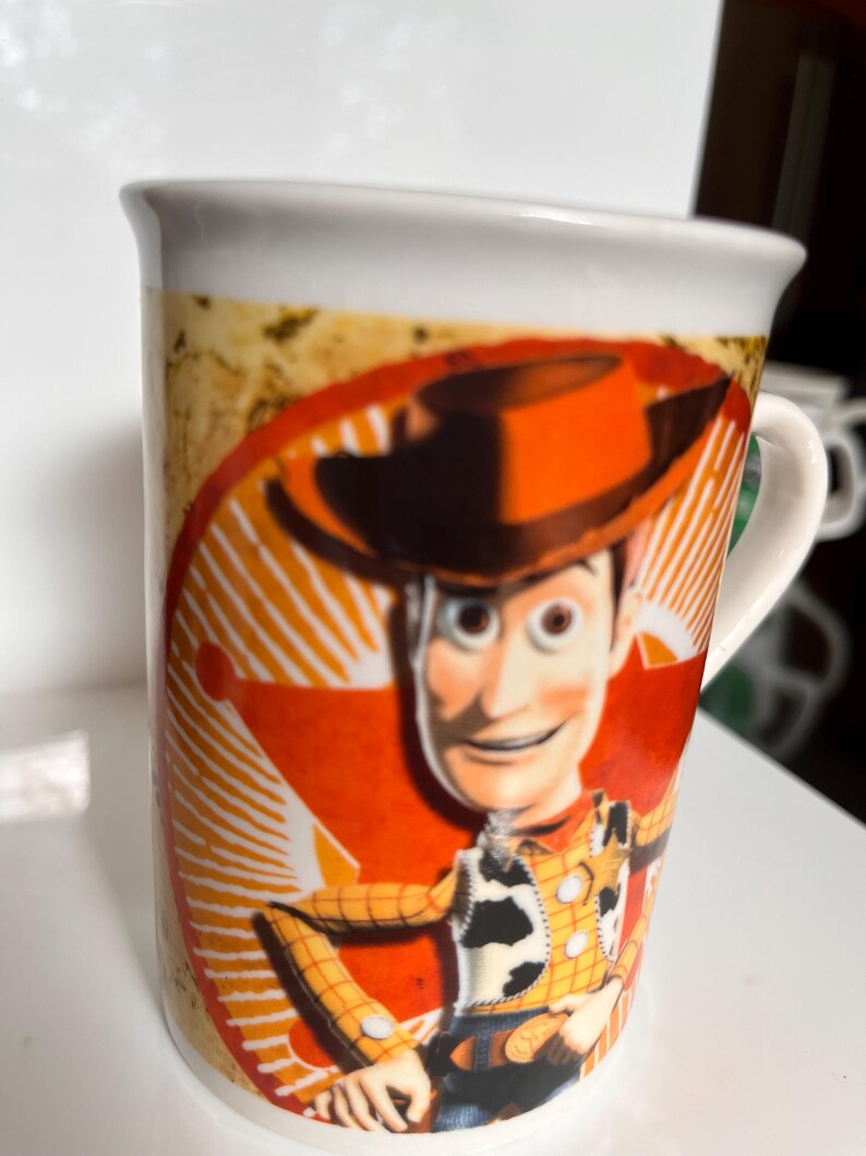 K&ouml;nnte beinhalten: Wei&szlig;er Keramikbecher mit einer farbenfrohen Illustration von Woody aus Toy Story. Das Design zeigt Woodys Gesicht, Hut und Outfit vor einem roten und orangefarbenen Hintergrund. Der Becher hat einen wei&szlig;en Henkel und Rand.