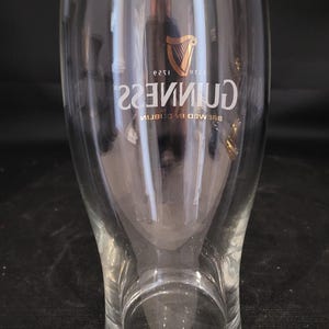May include: A clear, tulip-shaped Guinness pint glass. The glass features the Guinness logo in gold, with the harp symbol and the text "GUINNESS" in a curved font. The words "BREWED IN DUBLIN" are also visible.