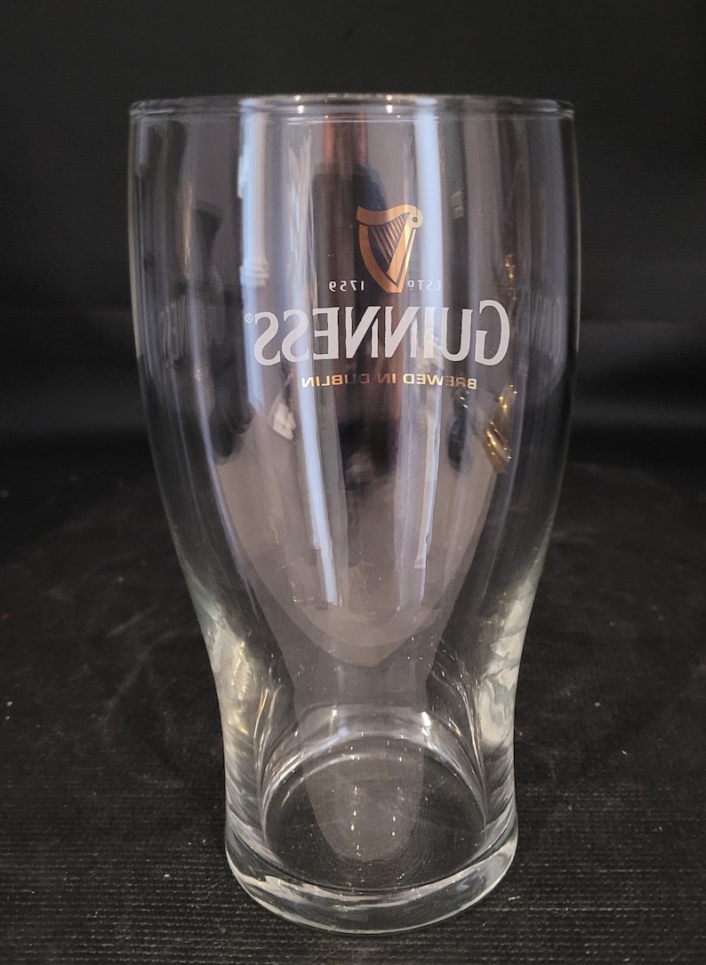 May include: A clear, tulip-shaped Guinness pint glass. The glass features the Guinness logo in gold, with the harp symbol and the text "GUINNESS" in a curved font. The words "BREWED IN DUBLIN" are also visible.