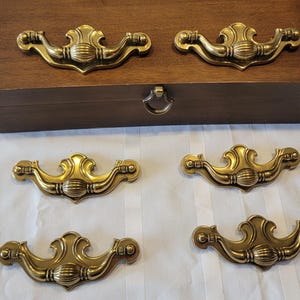 May include: A wooden box with a brass ring pull and six brass drawer pulls. The drawer pulls have an ornate design. The box and hardware are set against a white background.