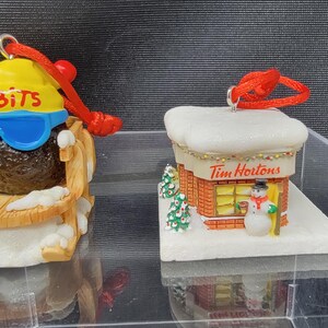 May include: Two Christmas ornaments. One is a Tim Hortons donut character wearing sunglasses and a yellow hat that says "Timbits". The other is a miniature Tim Hortons store with a snowman and Christmas trees.