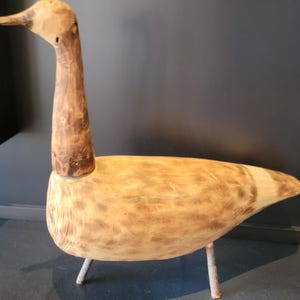 Peut inclure: Sculpture en bois en forme d'oie, avec un long cou et un corps arrondi. L'oie est faite de bois brun clair avec des accents brun foncé et repose sur trois fines pattes métalliques. L'œuvre a une esthétique rustique et naturelle.
