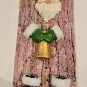May include: A festive wall hanging depicting Santa Claus's face, a gold bell, and boots with white trim. The background has a wood grain effect. The decoration is designed for the Christmas season.