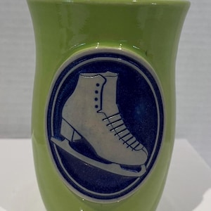 May include: A lime green ceramic mug featuring a central circular medallion. The medallion has a dark blue background with a raised, light-coloured relief of an ice skate. The mug has a smooth, glossy finish and a slightly flared top rim.