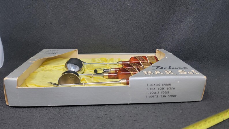 May include: A silver bar set in a box with a yellow lining. The set includes a mixing spoon, a cork screw, a double jigger, and a bottle can opener. The box is labeled "Deluxe Bar Set".