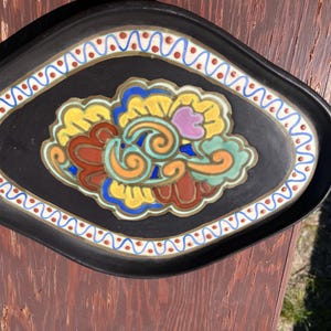 Puede incluir: Una bandeja ovalada de cerámica negra con un diseño floral central con detalles en esmalte amarillo, naranja, rojo, azul y morado claro. El borde está decorado con un patrón de ondas azules repetitivo y pequeños puntos rojos sobre fondo blanco.