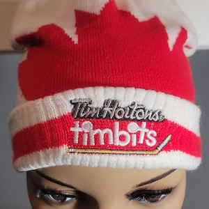 May include: A red and white knit beanie with a red pom-pom. The hat features a red maple leaf design and the Tim Hortons logo embroidered on the front. A winter hat.