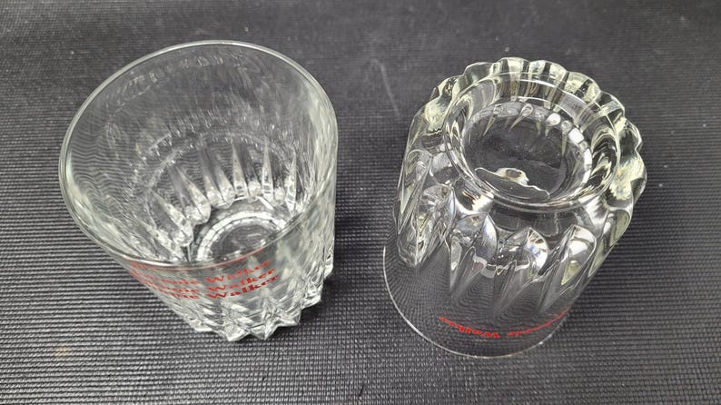 May include: Two clear, cut-glass tumblers with a diamond pattern and a scalloped rim. The text "Johnnie Walker" is printed in red near the base of each glass. The glasses are likely for serving whiskey or other spirits.