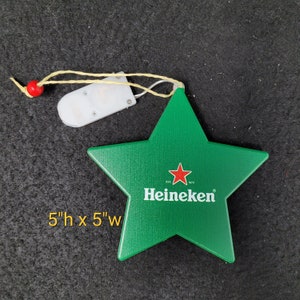 May include: Green star-shaped Heineken promotional item, 12.7 cm high and 12.7 cm wide, with the Heineken logo in white and a red star. It has a small white light and a string for hanging. The background is black.