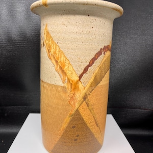 May include: A cylindrical ceramic vase featuring a speckled cream-coloured upper section and a solid tan lower section. The surface is decorated with intersecting diagonal bands in shades of golden yellow, deep brown, and rust-coloured speckles.