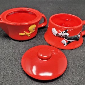 May include: A red ceramic teapot with a yellow bird design and a matching red ceramic mug with a black cat design. The teapot has a lid and a handle. The mug has a handle. There is also a red ceramic lid for the teapot.
