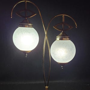 Könnte beinhalten: Eine Tischlampe aus Messing mit zwei weißen, mattierten Glaskugeln. Die Lampe hat ein einzigartiges Design mit geschwungenen Armen und augenförmigen Akzenten.