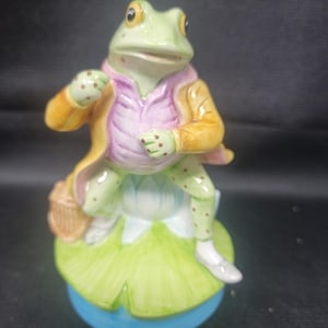 Puede incluir: Figura de cerámica de una rana vestida con un abrigo amarillo y una camisa rosa, sentada en una hoja de nenúfar verde. La rana tiene piel verde con lunares marrones y sostiene una pequeña cesta. La base es azul.