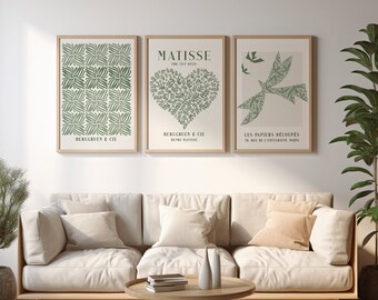 May include: Three framed art prints with botanical designs hang above a beige sofa. The prints feature green leaf patterns and text, including "MATISSE" and "BERGGRUEN & CIE." A round wooden coffee table and potted plants complete the living room scene.