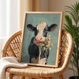 May include: A framed art print depicting a black and white cow holding flowers, set against a teal backdrop. The artwork is displayed on a wicker chair, with a cream-coloured blanket draped over it.