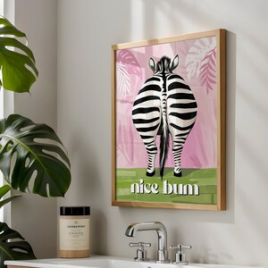 K&ouml;nnte beinhalten: Gerahmter Kunstdruck mit dem Hinterteil eines Zebras, mit schwarz-wei&szlig;en Streifen, vor einem rosa-gr&uuml;nen Hintergrund. Der Text "nice bum" ist in Wei&szlig; dargestellt. Das Kunstwerk befindet sich in einem hellen Holzrahmen.