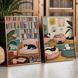 May include: Two framed art prints featuring cats. One print shows a black and white cat on a pink chair in a room with a bookshelf. The other print depicts several cats lounging on cushions. Both prints have gold frames.
