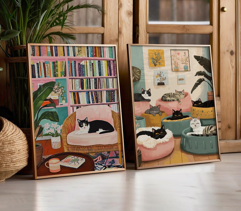 May include: Two framed art prints featuring cats. One print shows a black and white cat on a pink chair in a room with a bookshelf. The other print depicts several cats lounging on cushions. Both prints have gold frames.