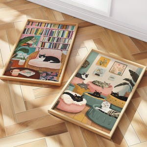 May include: Two framed art prints featuring cat illustrations. One print depicts a cat lounging in a chair in a room with a bookshelf. The other shows multiple cats in beds. Both prints have wooden frames.