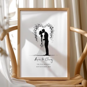 May include: A framed print with a silhouette of a kissing couple beneath a floral heart. The names "Ann & Chay" and the date "16th November 2025" are written below. The frame is light brown, and the print is displayed on a wooden chair.
