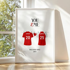 May include: A framed print with the text "YOU & ME". Below, two red football shirts with white numbers and names hang from a washing line. A red heart is between the shirts. The text "TOGETHER SINCE 2021" is at the bottom of the print.