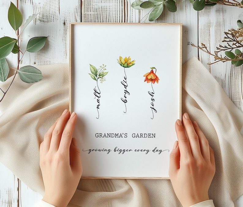 May include: A framed print with a white background and three watercolor flowers. The flowers are labeled "Josh", "Brydie", and "Matt". The text below the flowers reads "Grandma's Garden growing bigger every day".