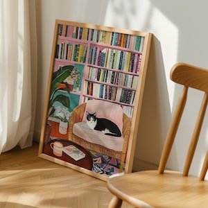 May include: A framed art print depicts a cozy interior scene. A black and white cat rests on a pink cushion in a wicker chair. A bookshelf filled with books is in the background. A small table with a mug and book is in front of the chair.