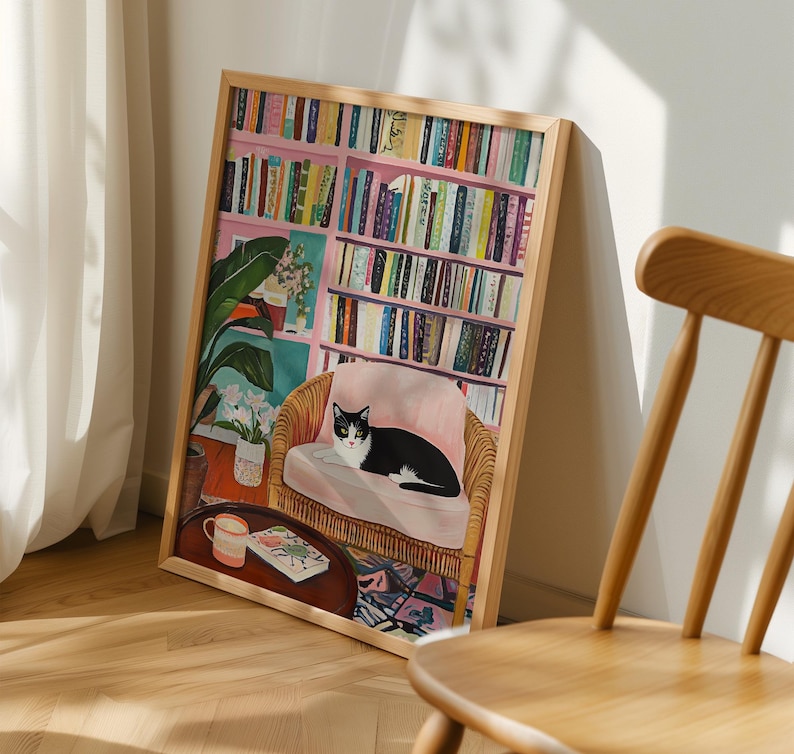 May include: A framed art print depicts a cozy interior scene. A black and white cat rests on a pink cushion in a wicker chair. A bookshelf filled with books is in the background. A small table with a mug and book is in front of the chair.