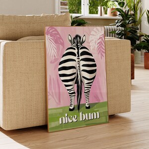 K&ouml;nnte beinhalten: Ein gerahmter Kunstdruck mit dem Hinterteil eines Zebras mit schwarz-wei&szlig;en Streifen, vor einem rosa Hintergrund mit tropischen Bl&auml;ttern. Der Schriftzug "nice bum" steht in Gr&uuml;n am unteren Rand.