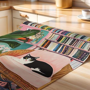 May include: A colorful, artistic table runner featuring a black and white cat lounging on a pink cushion. The runner showcases a painted scene with bookshelves, plants, and a wicker chair. The color palette includes pink, green, and various book colors.