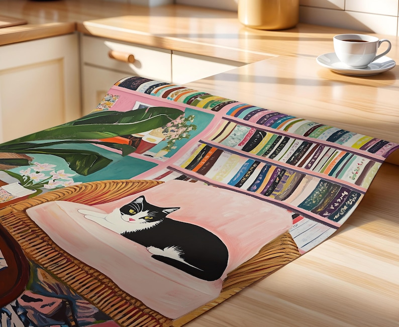 May include: A colorful, artistic table runner featuring a black and white cat lounging on a pink cushion. The runner showcases a painted scene with bookshelves, plants, and a wicker chair. The color palette includes pink, green, and various book colors.