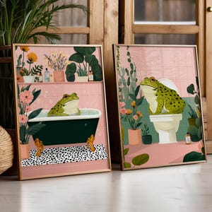 Può includere: Due stampe artistiche incorniciate. Una raffigura una rana verde in una vasca da bagno, l'altra una rana su un water. Entrambe le stampe hanno uno sfondo di piastrelle rosa con piante e fiori. Le cornici sono di colore legno chiaro.