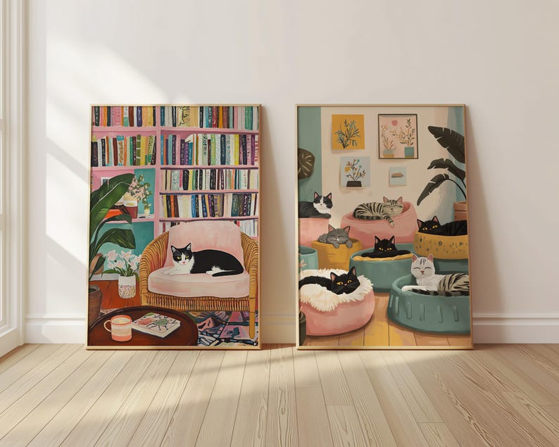 May include: Two framed art prints featuring cats. One shows a black and white cat on a pink chair in front of a bookshelf. The other depicts multiple cats lounging on cushions. Both have gold frames.