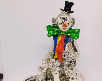 May include: A silver figurine of a clown riding a bicycle. The clown is wearing a black top hat, a green and white polka dot bow tie, and a red, yellow, and blue striped shirt. The clown has a red nose and a painted-on smile.