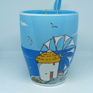 May include: A light blue ceramic mug with a matching spoon. The mug features a seaside illustration with a windmill, seagulls, and a blue sky. The spoon rests on the rim, held by a gold wire.