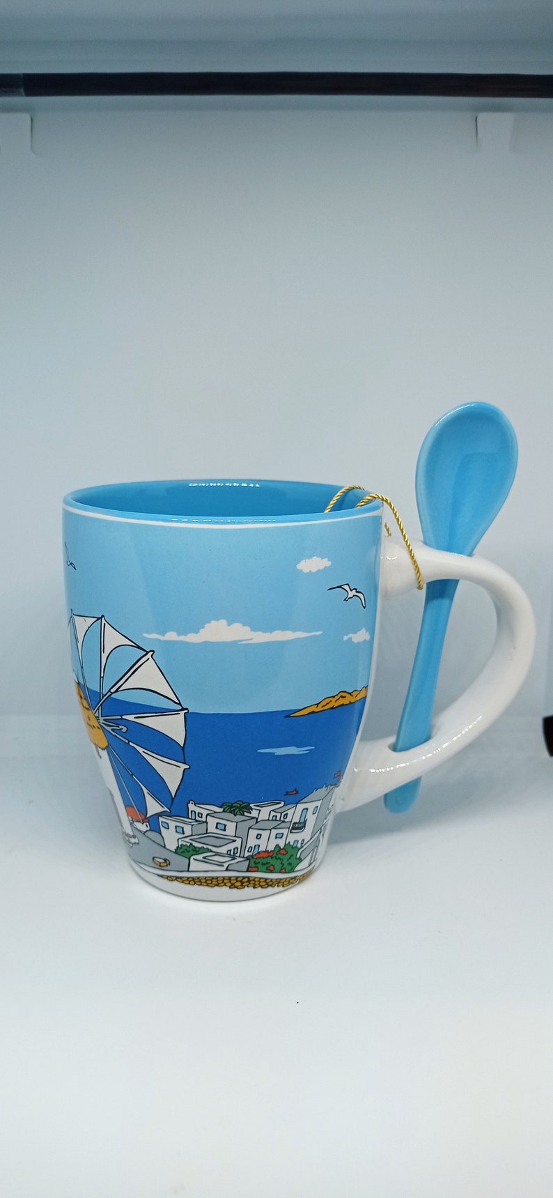 May include: A ceramic mug with a light blue interior and exterior, featuring a seaside illustration with a windmill, buildings, and a blue spoon attached to the handle. The mug has a white handle and a gold string.