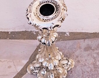 May include: A silver candlestick with an ornate design. The candlestick has a round base with a decorative pattern and a tall, slender stem.