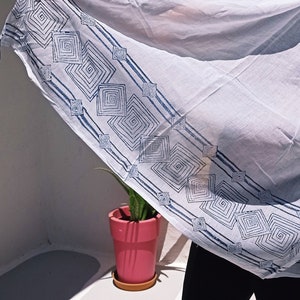 May include: A white cotton scarf with a blue geometric pattern. The scarf is being held by a person wearing black pants.