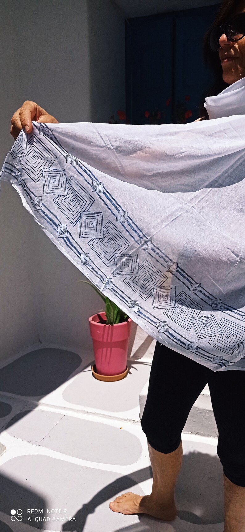 May include: A white cotton scarf with a blue geometric pattern. The scarf is being held by a person wearing black pants.